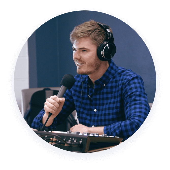 A person wearing headphones and a blue plaid shirt speaks into a microphone, sitting at a desk with audio equipment.